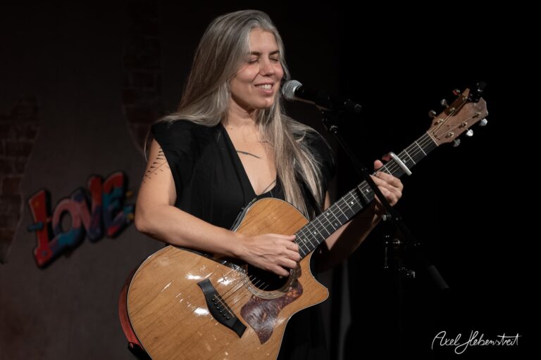 Eine Musikerin spielt mit Leidenschaft Gitarre und singt auf einer Bühnen, umgeben von einer lebhaften Kulisse. – © Axel Hebenstreit