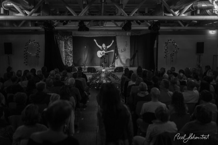 Eine Künstlerin mit Gitarre begeistert das Publikum in einem gemütlichen Konzertsaal – © Axel Hebenstreit