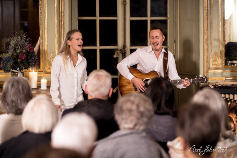 Zwei Musiker singen und spielen Gitarre vor einem Publikum in einem eleganten Ambiente. – © Axel Hebenstreit