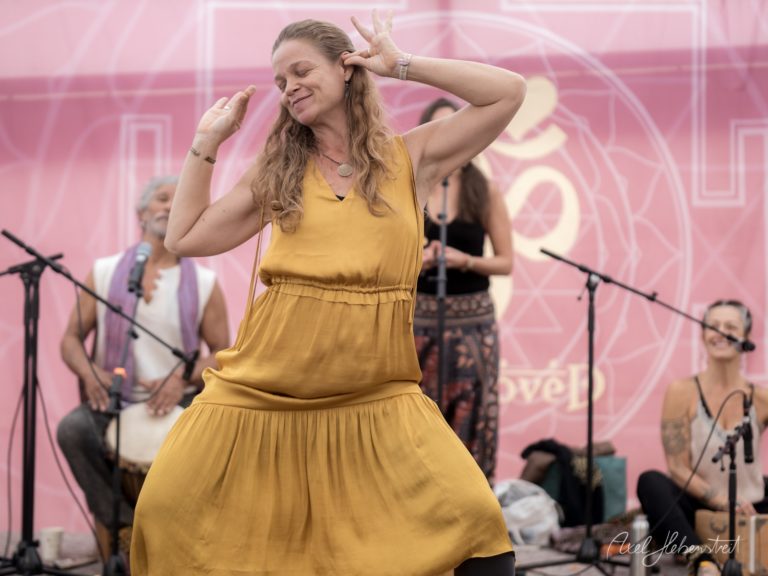 Frau in gelbem Kleid tanzt freudig auf einer Bühne mit rosa Hintergrund vor Publikum und Band – © Axel Hebenstreit