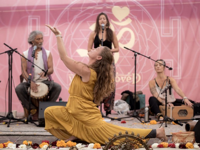 Frau in goldenem Kleid singt kniend bei spirituellem Konzert mit Live-Musikern und Blütendekor vor rosa Bühne – © Axel Hebenstreit