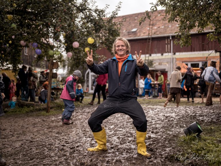 Glücklicher Mann springt mit Peace-Zeichen in gelbem Stiefeln auf Dorfplatz mit feiernden Menschen und Häusern im Hintergrund – © Axel Hebenstreit