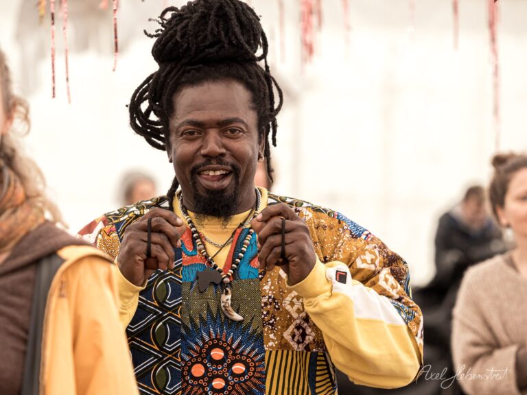 Lachender Mann mit Dreadlocks in farbenfrohem afrikanischem Gewand und Schmuck auf Festival – © Axel Hebenstreit