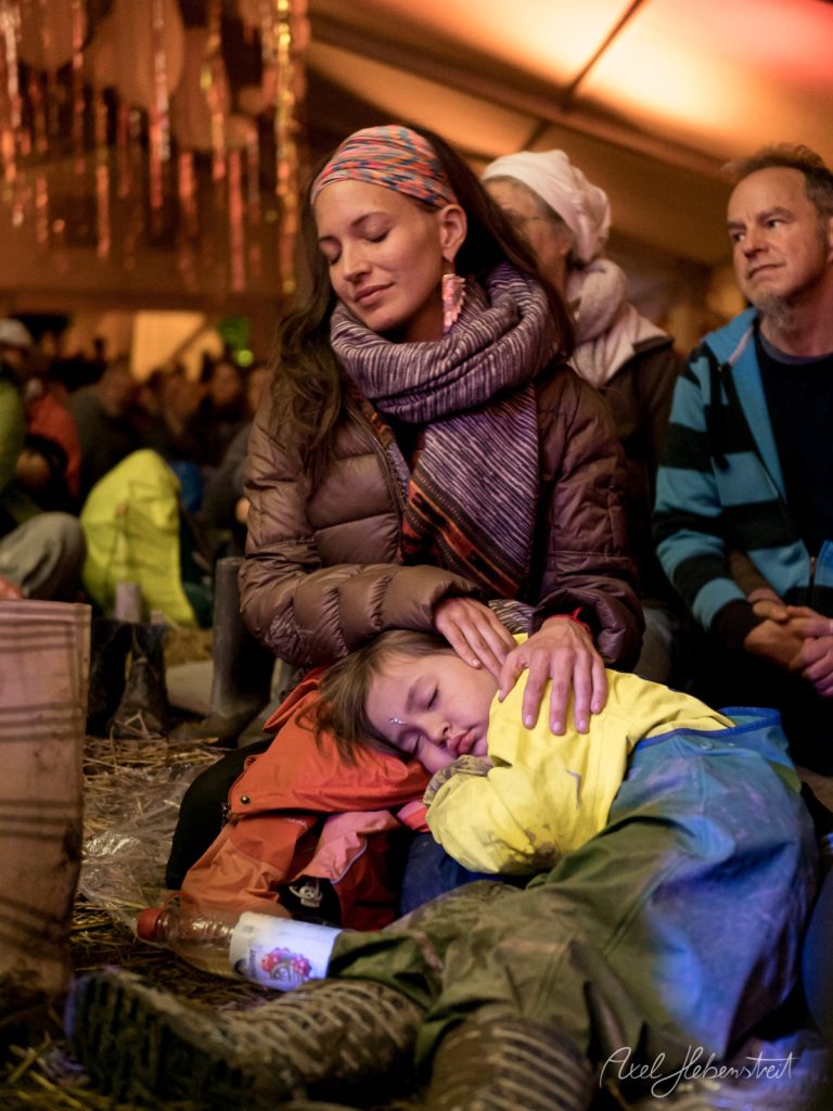 Mutter beruhigt ihr schlafendes Kind in warmer Zeltumgebung während eines Festivals – © Axel Hebenstreit