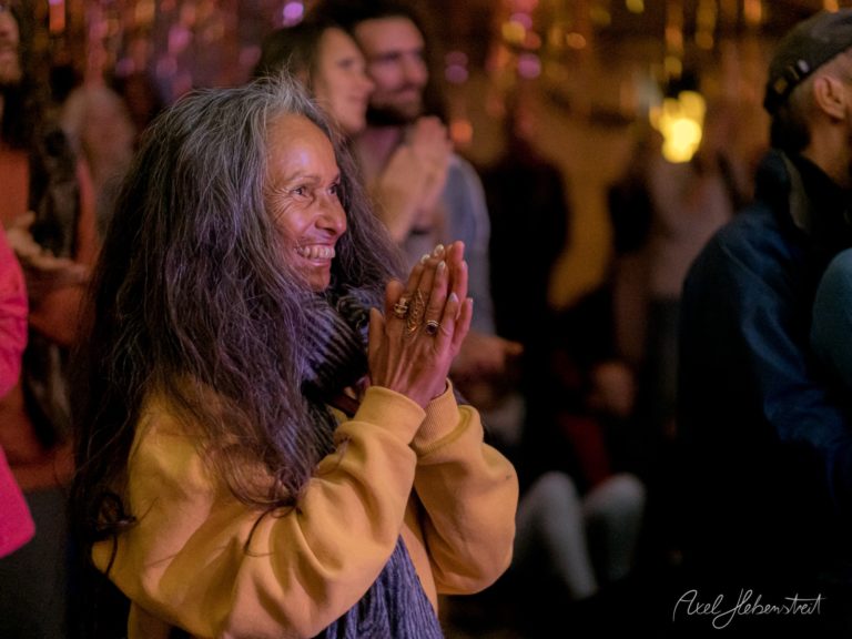 Frau mit langen Haaren in gelbem Pullover klatscht begeistert auf Konzert oder Festival mit farbiger Bühnenbeleuchtung – © Axel Hebenstreit