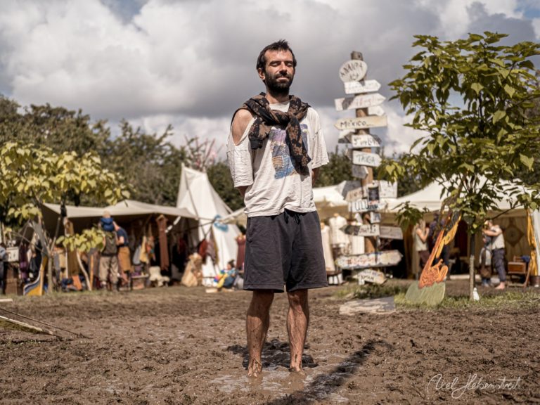 Mann mit Bart steht in Pfütze auf Festivalgelände mit Zelten und Schildern im Hintergrund – © Axel Hebenstreit