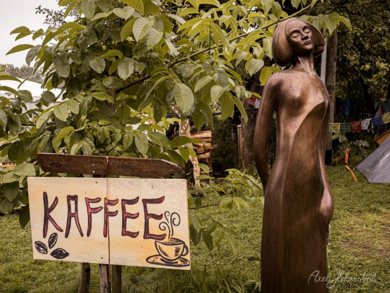 Bronzeskulptur einer Frau neben handgemaltem Kaffee-Schild in grünem Garten – © Axel Hebenstreit