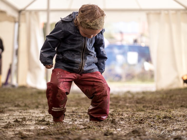 Kleines Kind in Regenjacke und roten Hosen spielt ausgelassen im Schlamm unter einem Festivalzelt – © Axel Hebenstreit