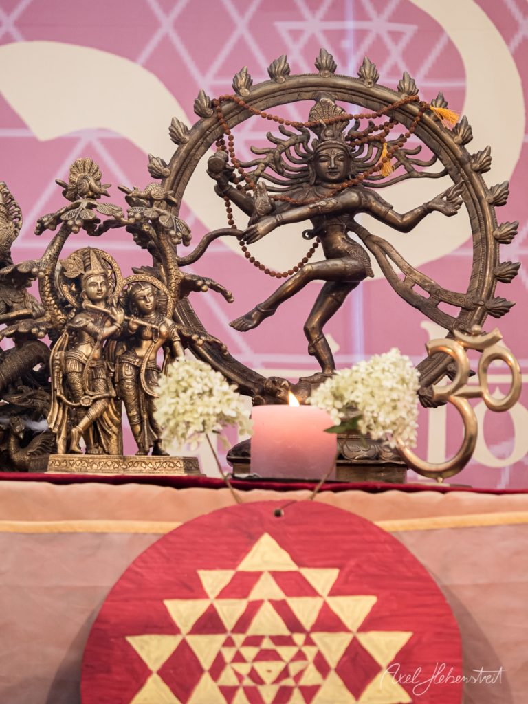 Bronzefiguren von Shiva und Krishna in Tanzpose vor rosa Hintergrund mit Blumen und Kerze – © Axel Hebenstreit