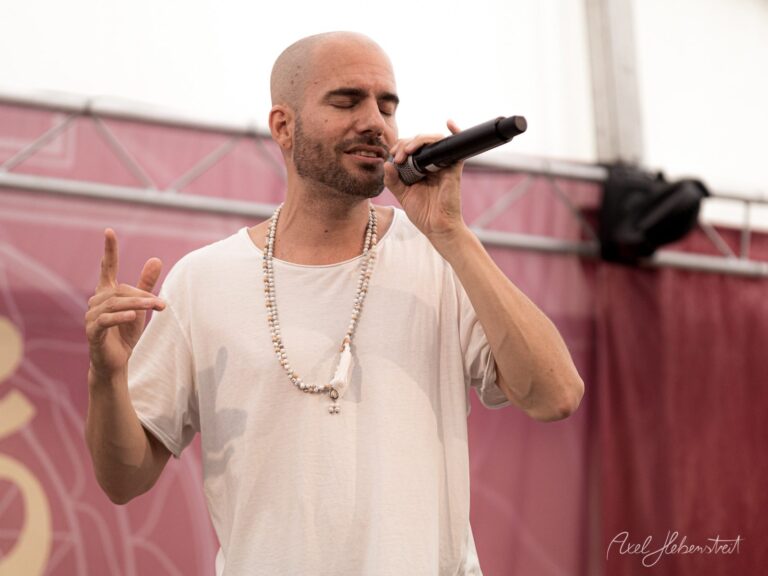 Sänger mit Mikrofon und Perlenkette performt auf Festival-Bühne vor rosa Hintergrund – © Axel Hebenstreit