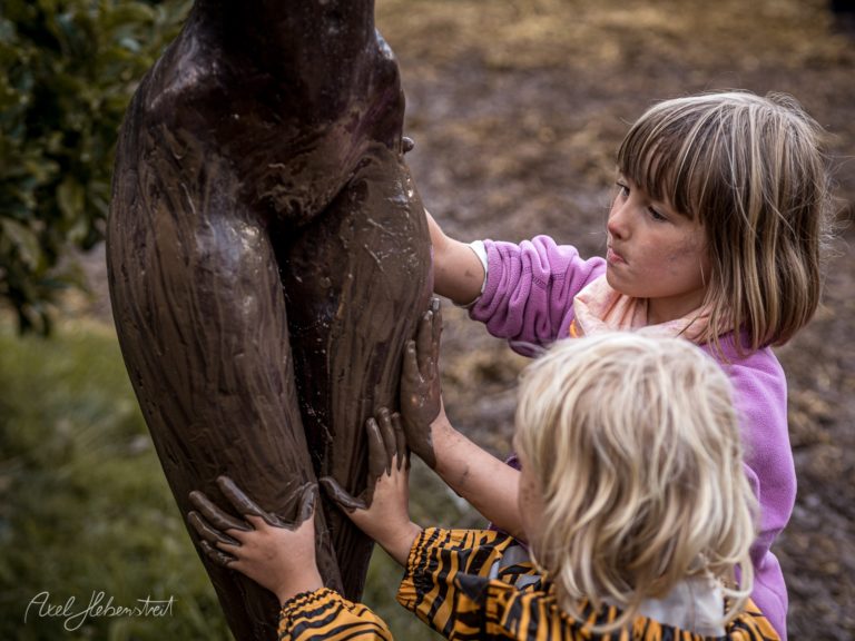 Zwei Kinder erkunden neugierig eine dunkle Holzskulptur in der Natur – © Axel Hebenstreit