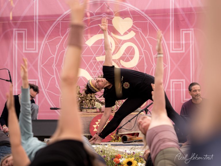 Yoga-Lehrer in schwarzer Kleidung führt eine Rückbeuge-Pose vor rosa illuminiertem Mandala-Hintergrund mit Zuschauern im Vordergrund – © Axel Hebenstreit