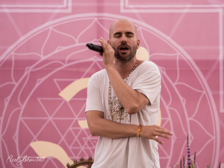 Ein Mann singt leidenschaftlich in ein Mikrofon vor einem bunten, geometrisch gestalteten Hintergrund – © Axel Hebenstreit