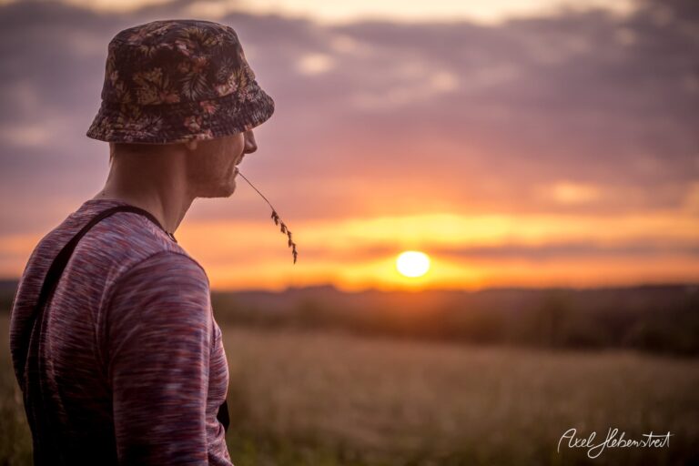 Mann mit Sonnenhut und Halm im Mund beobachtet den Sonnenuntergang in einer ländlichen Umgebung – © Axel Hebenstreit