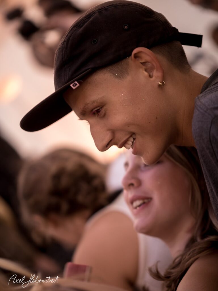 Junger Mann mit Kappe und funkelndem Gesichtsausdruck bei einem Festival, lächelnd im Gespräch mit einer Freundin. – © Axel Hebenstreit