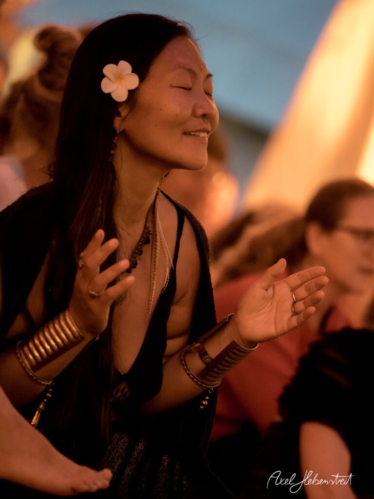 Eine Frau mit einer Blume im Haar meditiert begeistert, umgeben von einer warmen Festivalatmosphäre – © Axel Hebenstreit