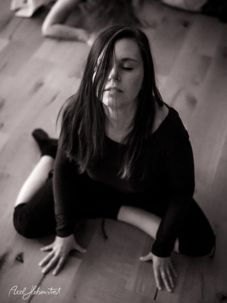 Frau in meditativer Pose auf dem Boden, umgeben von sanfter Lichtstimmung und holziger Textur – © Axel Hebenstreit