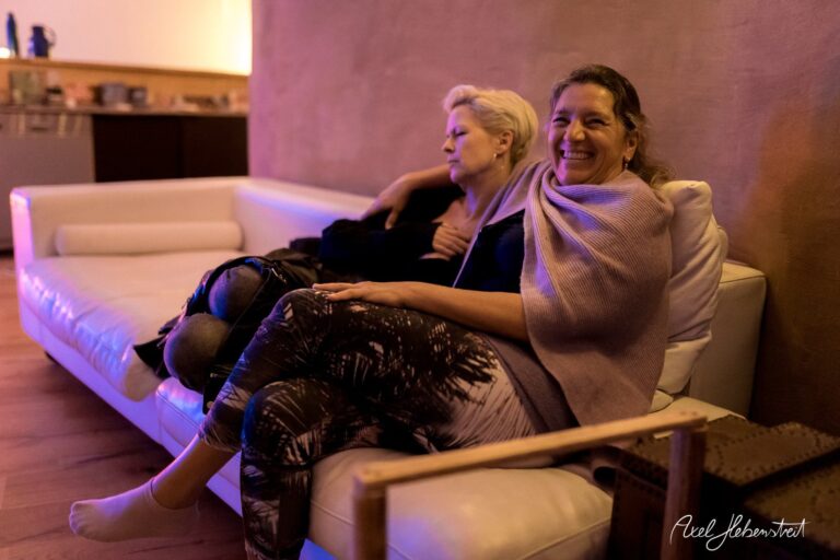Zwei Frauen sitzen entspannt auf einer Couch in einem warm beleuchteten Raum und lachen miteinander. – © Axel Hebenstreit
