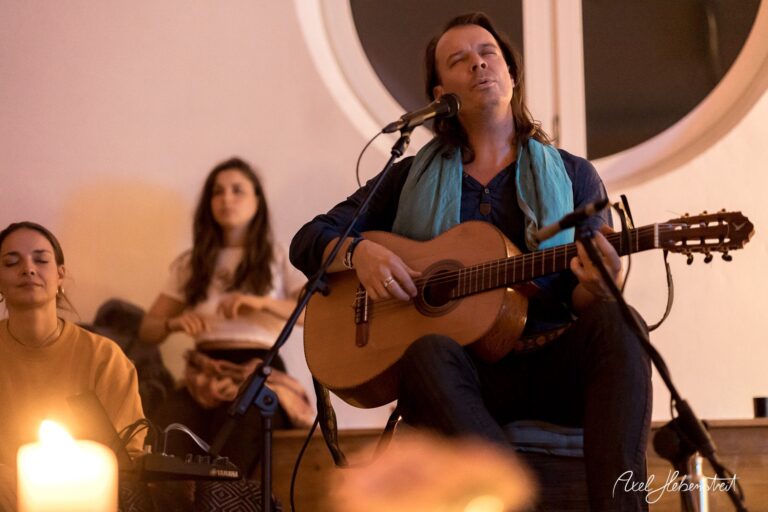 Ein Singer-Songwriter sitzt mit Gitarre auf der Bühne und singt, während Zuhörer entspannt lauschen – © Axel Hebenstreit