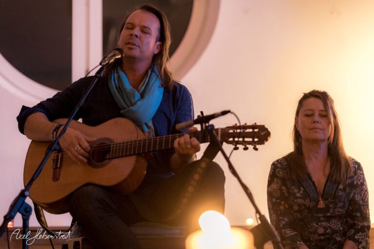 Ein Sänger mit Gitarre auf einer spirituellen Veranstaltung, während eine Frau meditiert – © Axel Hebenstreit