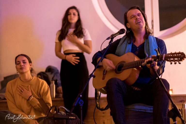 Ein Musiker spielt Gitarre, während zwei Zuhörer in Meditation versunken sind, in einem stimmungsvollen Raum. – © Axel Hebenstreit