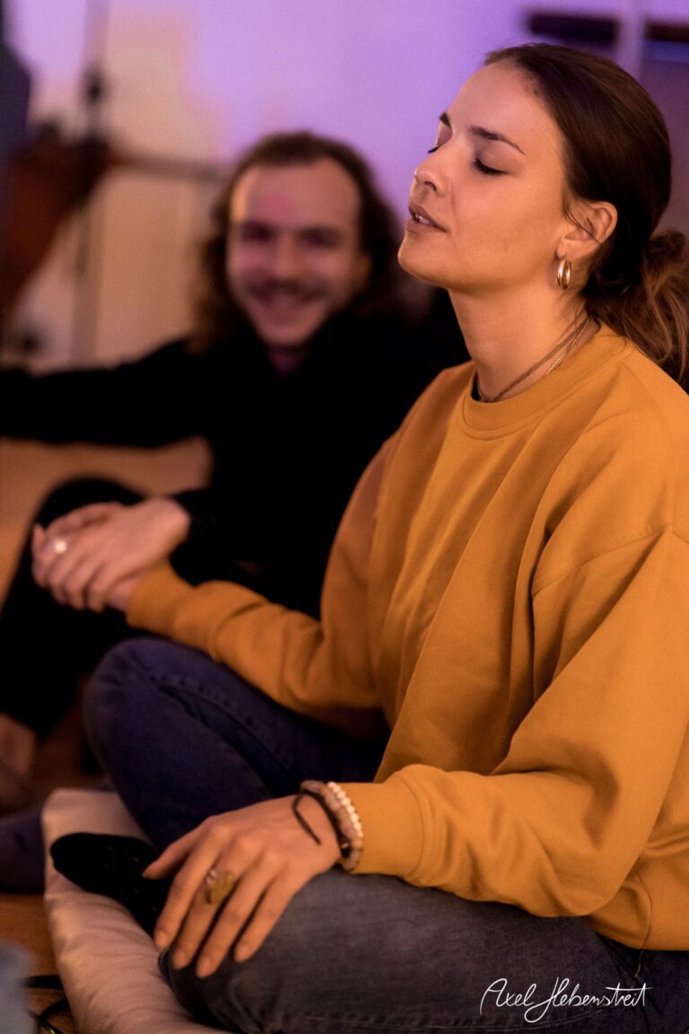 Eine entspannte Frau in einem orangefarbenen Sweatshirt meditiert während einer Gruppenaktivität – © Axel Hebenstreit.