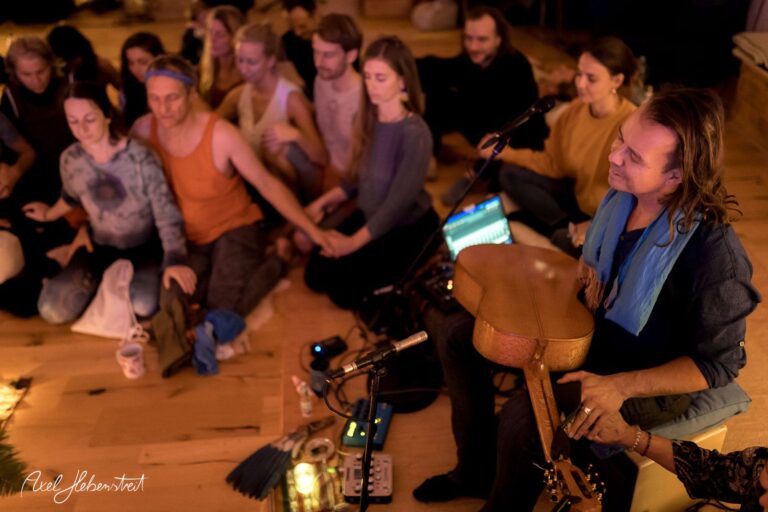 Eine Gruppe von Menschen sitzt um einen Musiker mit Gitarre und lauscht der meditativen Musik in einem warmen Raum – © Axel Hebenstreit