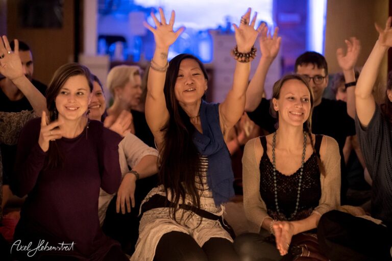 Frauen und Männer sitzen umgeben von Menschen und heben begeistert die Hände in einer festlichen Yoga-Atmosphäre – © Axel Hebenstreit