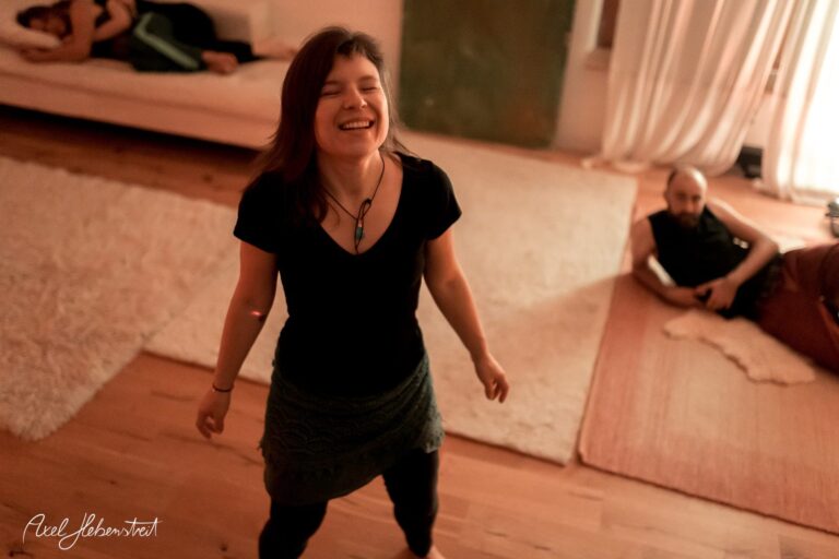 Lächelnde Frau, die glücklich tanzt, während eine andere Person im Hintergrund entspannt auf dem Boden sitzt – © Axel Hebenstreit