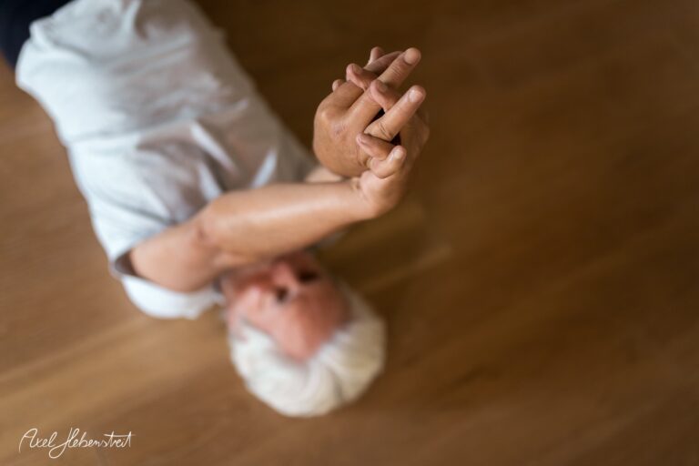 Eine ältere Person stretchend auf einem Holzfußboden, das Hauptaugenmerk liegt auf den Händen. – © Axel Hebenstreit