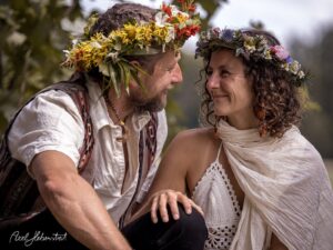 Ein Paar mit Blumenkränzen lächelt sich in einer natürlichen Umgebung liebevoll an – © Axel Hebenstreit