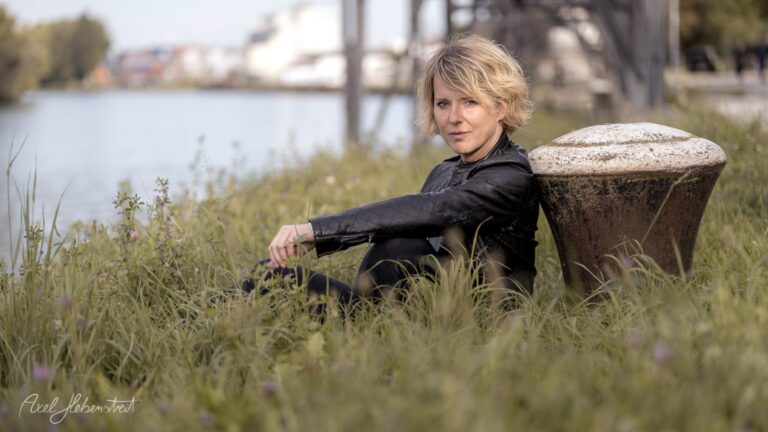 Frau sitzt elegant im hohen Gras am Ufer eines Kanals und blickt nachdenklich in die Kamera – © Axel Hebenstreit