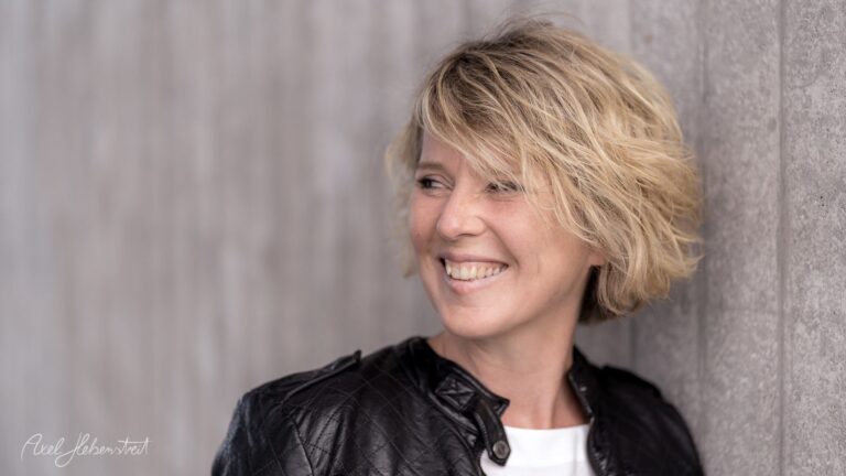 Lächelnde Frau mit lockigem Haar vor einer grauen Wand, die eine positive und entspannte Stimmung ausstrahlt – © Axel Hebenstreit.