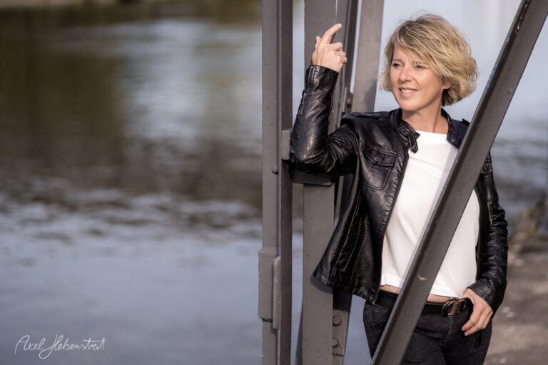 Eine lächelnde Frau in einer Lederjacke lehnt entspannt an einer Metallstruktur am Wasser. – © Axel Hebenstreit