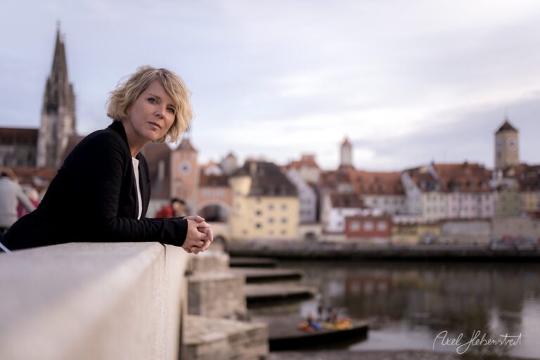 Eine Frau mit kurzer Frisur blickt nachdenklich über die Brücke auf die Altstadt von Regensburg – © Axel Hebenstreit