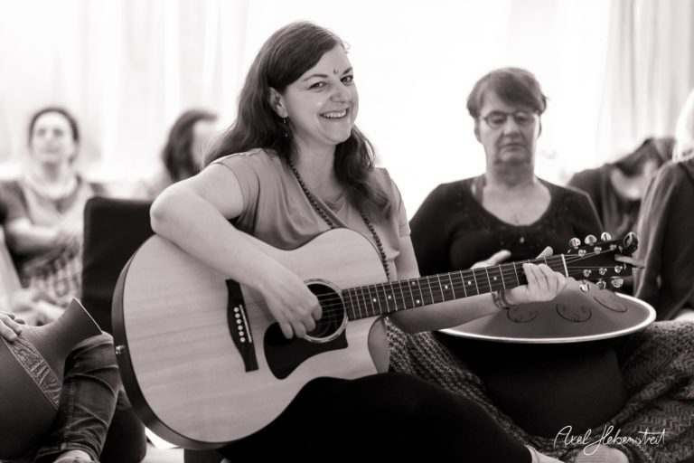 Eine lächelnde Frau spielt Gitarre in einer harmonischen Runde von Menschen, die entspannt zuhören. – © Axel Hebenstreit