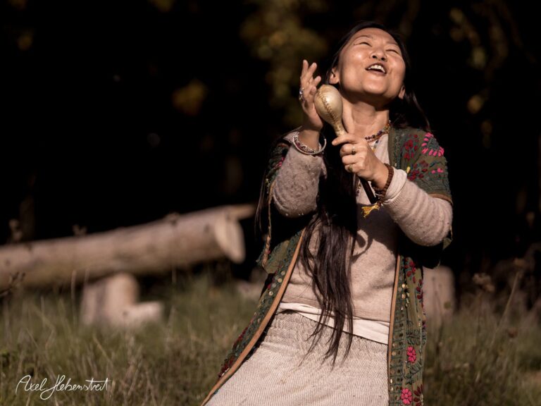 Frau in traditioneller Kleidung, die fröhlich bei Sonnenlicht eine Schüttelriemen spielt – © Axel Hebenstreit