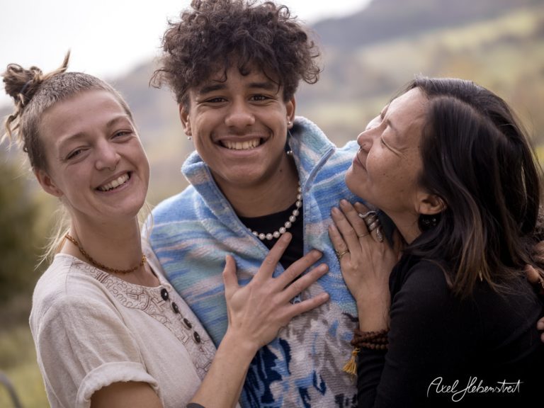 Drei junge Menschen lachen und umarmen sich vor einer malerischen Landschaft. – © Axel Hebenstreit