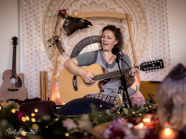 Frau mit Gitarre in harmonischer Umgebung singt und spielt, umgeben von Blumen und sanfter Beleuchtung – © Axel Hebenstreit