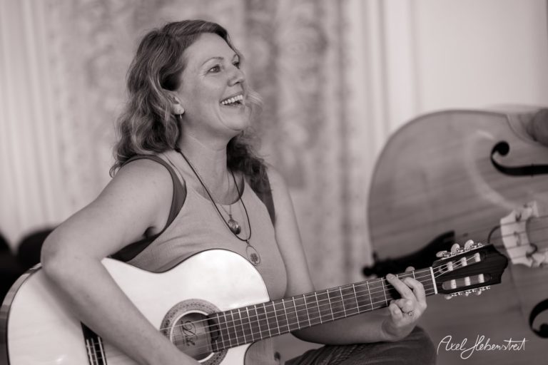 Frau mit Gitarre lächelt fröhlich, während sie in einer gemütlichen Umgebung musiziert – © Axel Hebenstreit