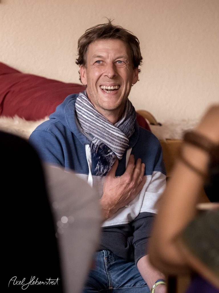 Lächender Mann in blauer Jacke und Schal, der in entspannter Runde interessiert lauscht – © Axel Hebenstreit