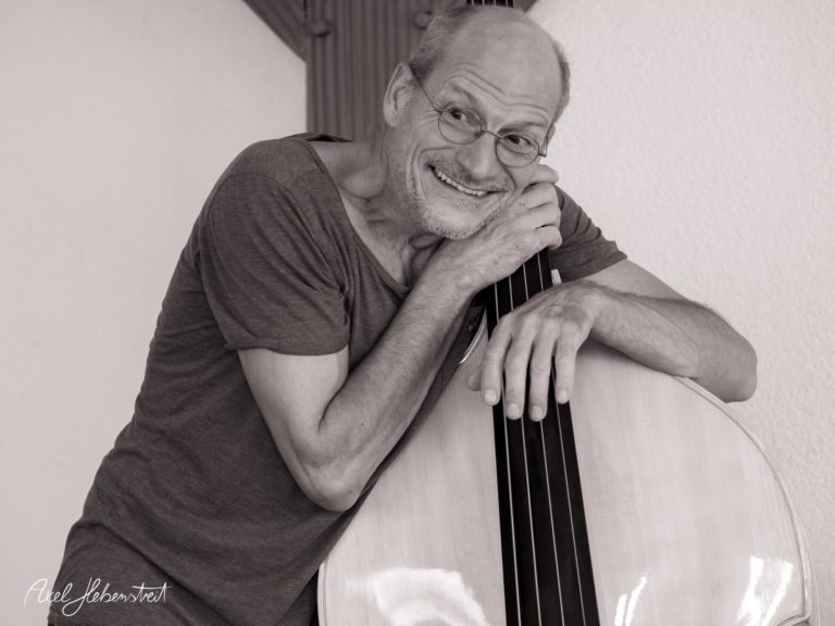 Lächelnder Musiker umarmt einen Kontrabass in einem hellen, freundlichen Raum – © Axel Hebenstreit
