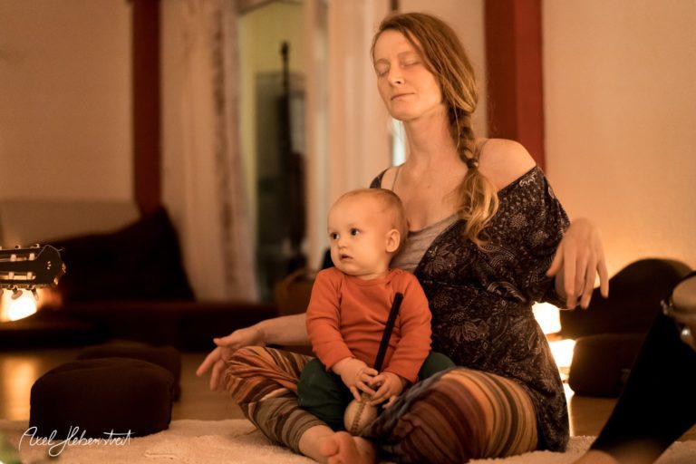 Eine Mutter meditiert entspannt im Wohnzimmer, während ihr Kleinkind neben ihr sitzt und spielt – © Axel Hebenstreit