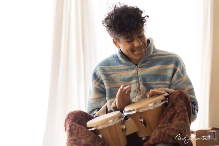 Ein junger Mann spielt entspannt auf Bongos in einer hellen, gemütlichen Umgebung – © Axel Hebenstreit