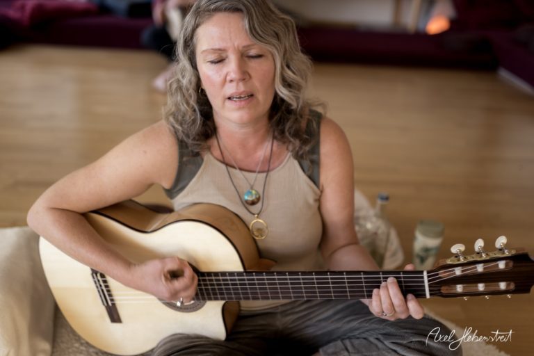 Eine Frau spielt konzentriert Gitarre und singt in einem hellen Raum mit Holzfußboden – © Axel Hebenstreit