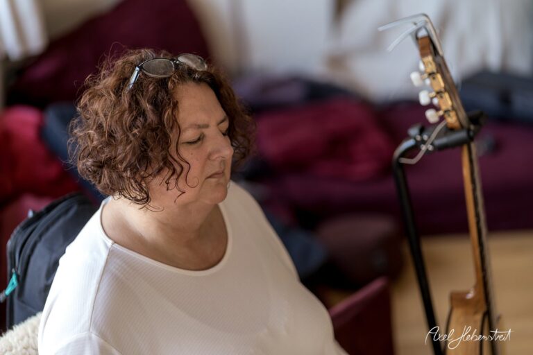 Frau mit lockigem Haar sitzt nachdenklich in einem Raum mit einer Gitarre im Hintergrund – © Axel Hebenstreit