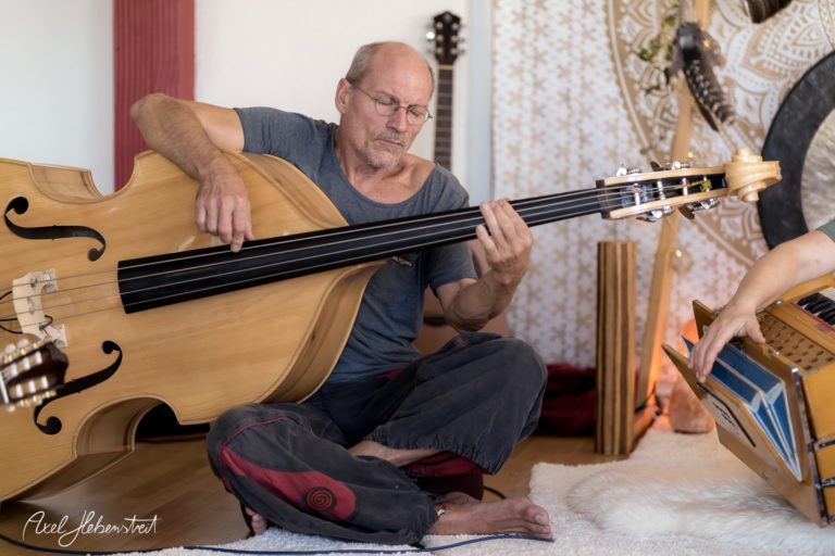 Ein Musiker sitzt entspannt im Schneidersitz und spielt auf einem großen, handgefertigten Musikinstrument – © Axel Hebenstreit.