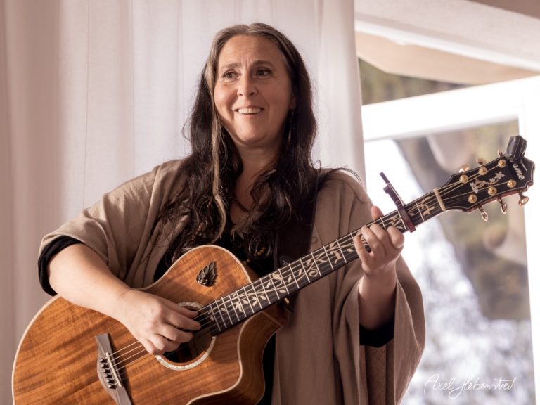 Eine lächelnde Musikerin mit Gitarre steht vor einem Fenster und spielt in einer entspannten Atmosphäre. – © Axel Hebenstreit