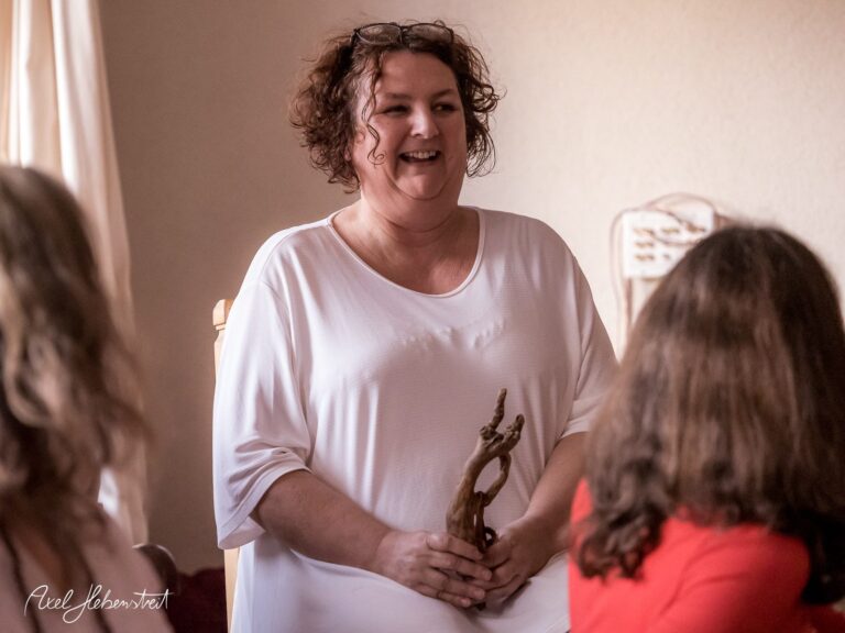 Eine lächelnde Frau in einem weißen Kleid sitzt entspannt und hält einen Holzgegenstand – © Axel Hebenstreit