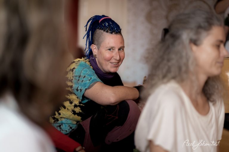 Frau mit blauen Zöpfen lächelt in einer entspannten Atmosphäre, umgeben von anderen Personen bei einem Event – © Axel Hebenstreit.